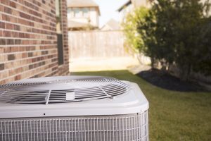 An air conditioner unit outside a brick home in a residential neighborhood. The air conditioner is in a back yard in the hot summer season