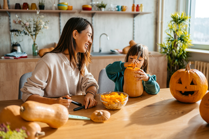 Young mother and daughter sitting in the kitchen and carving out pumpkins for Halloween.